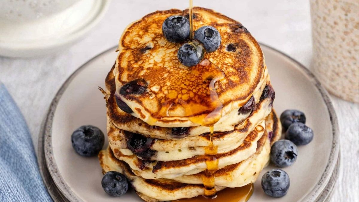 Stack of fluffy blueberry pancakes topped with fresh blueberries as maple syrup is poured over the top and drips down the sides.