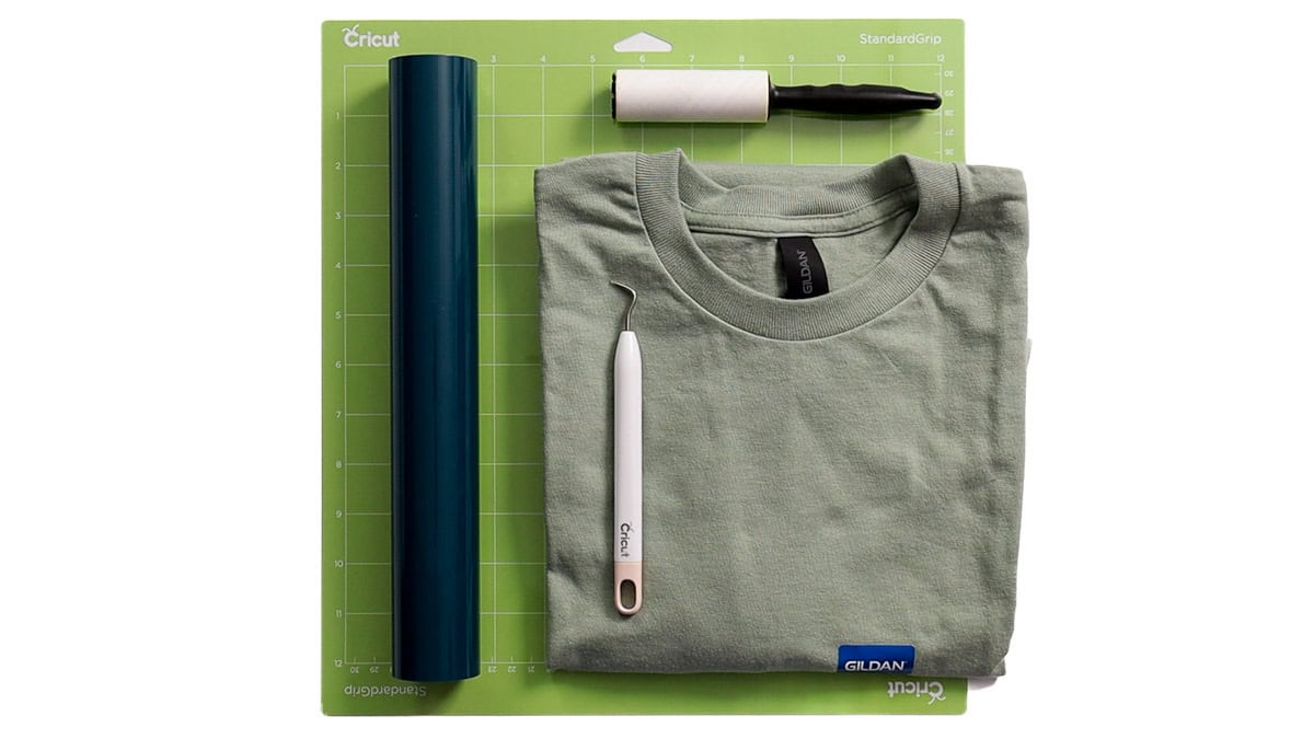 Flat lay of Cricut shirt-making supplies including a green T-shirt, dark blue iron-on vinyl, weeding tool, lint roller, and Cricut cutting mat.