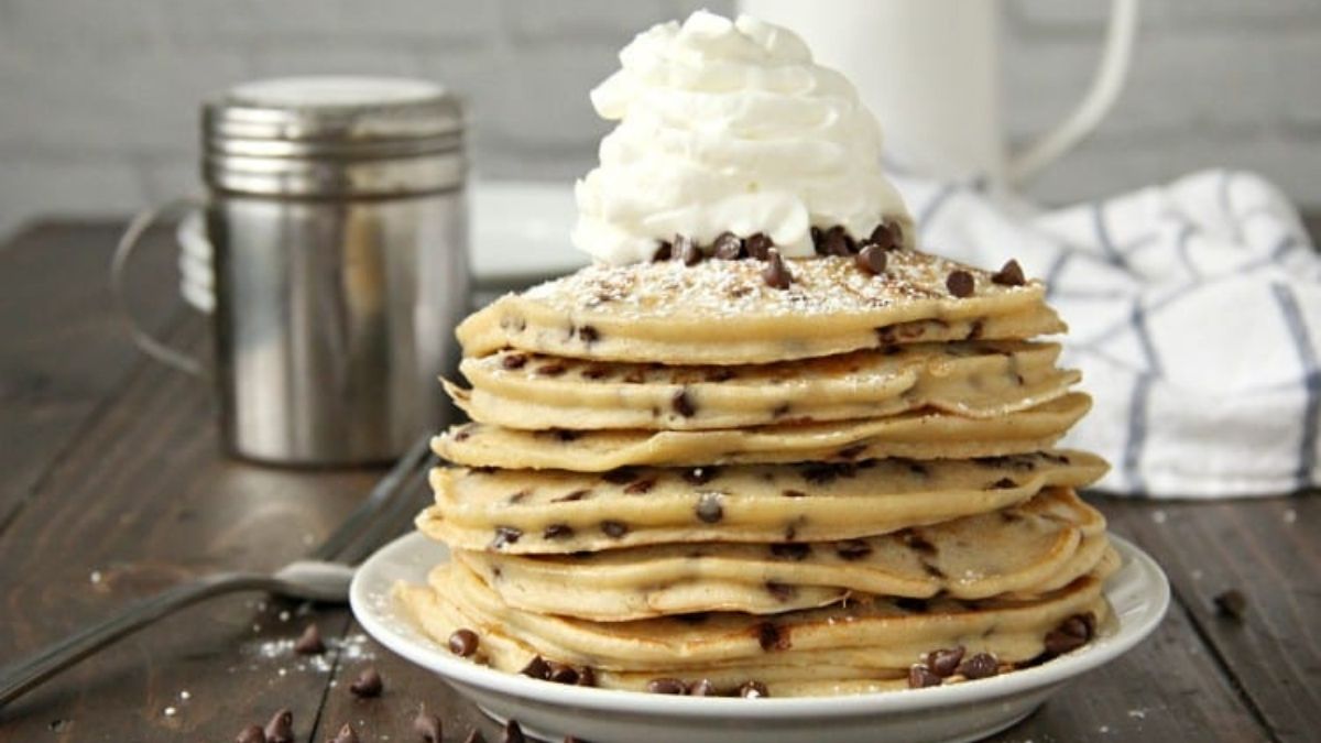 Stack of cannoli pancakes filled with chocolate chips and topped with whipped cream, mini chocolate chips, and powdered sugar on a white plate.
