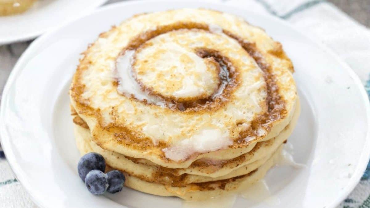 Stack of cinnamon swirl pancakes drizzled with sweet icing and topped with a cinnamon sugar spiral, served on a white plate with blueberries.