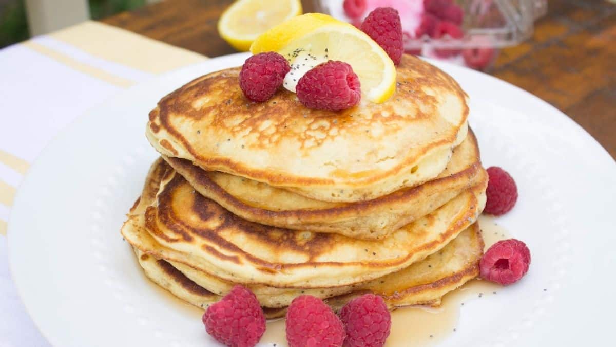 Stack of fluffy lemon poppy seed pancakes topped with fresh raspberries, lemon slices, and syrup on a white plate.