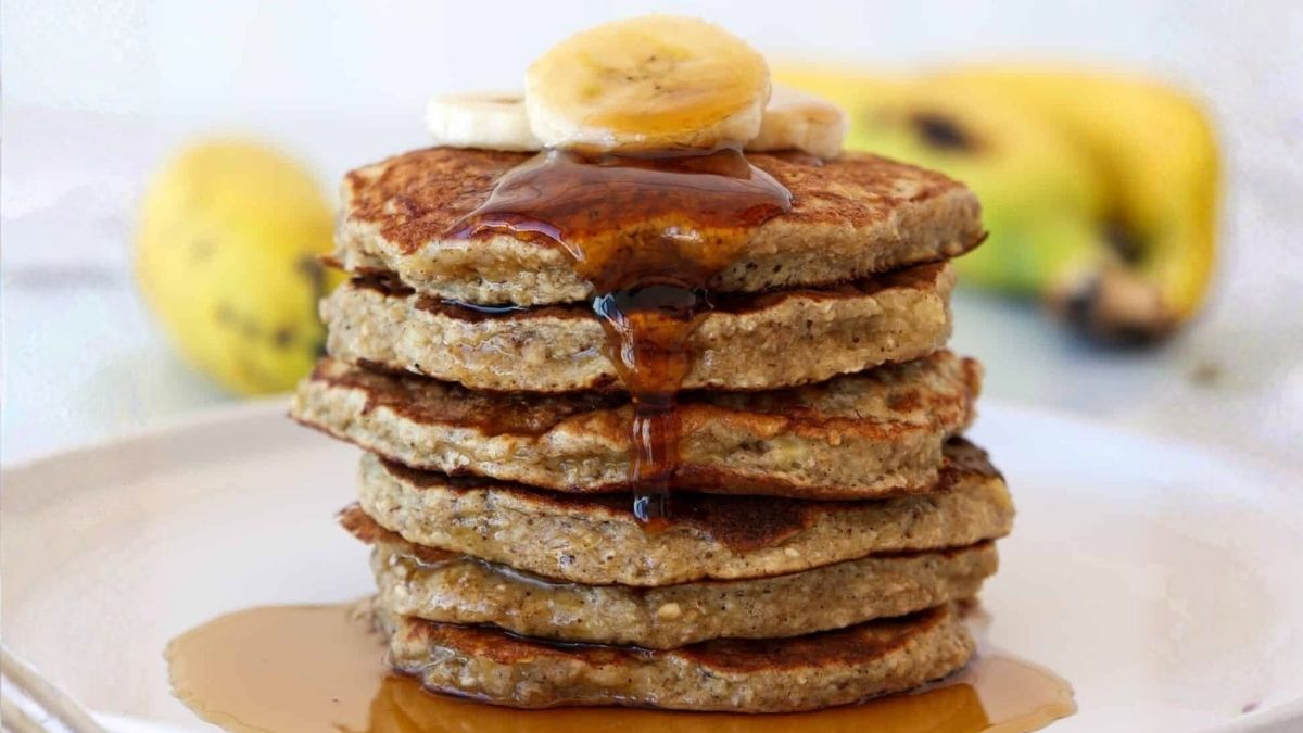 Stack of healthy banana oatmeal pancakes topped with banana slices and maple syrup, served on a white plate with fresh bananas in the background.