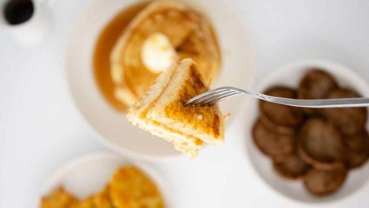 Fork holding a bite of fluffy pancake in focus, with a stack of pancakes and a bowl of sausage patties blurred in the background.