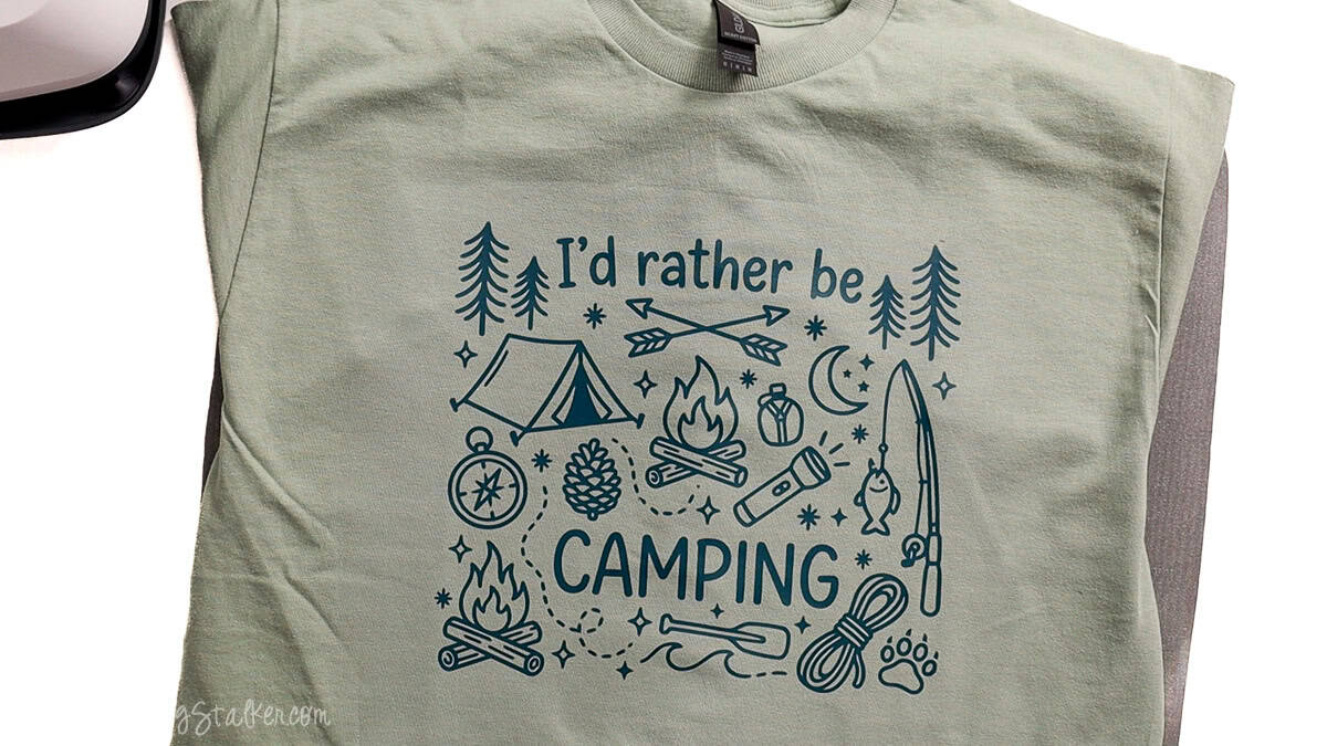 Light green T-shirt featuring an “I’d Rather Be Camping” design with a tent, campfire, trees, fishing pole, flashlight, and outdoor icons.