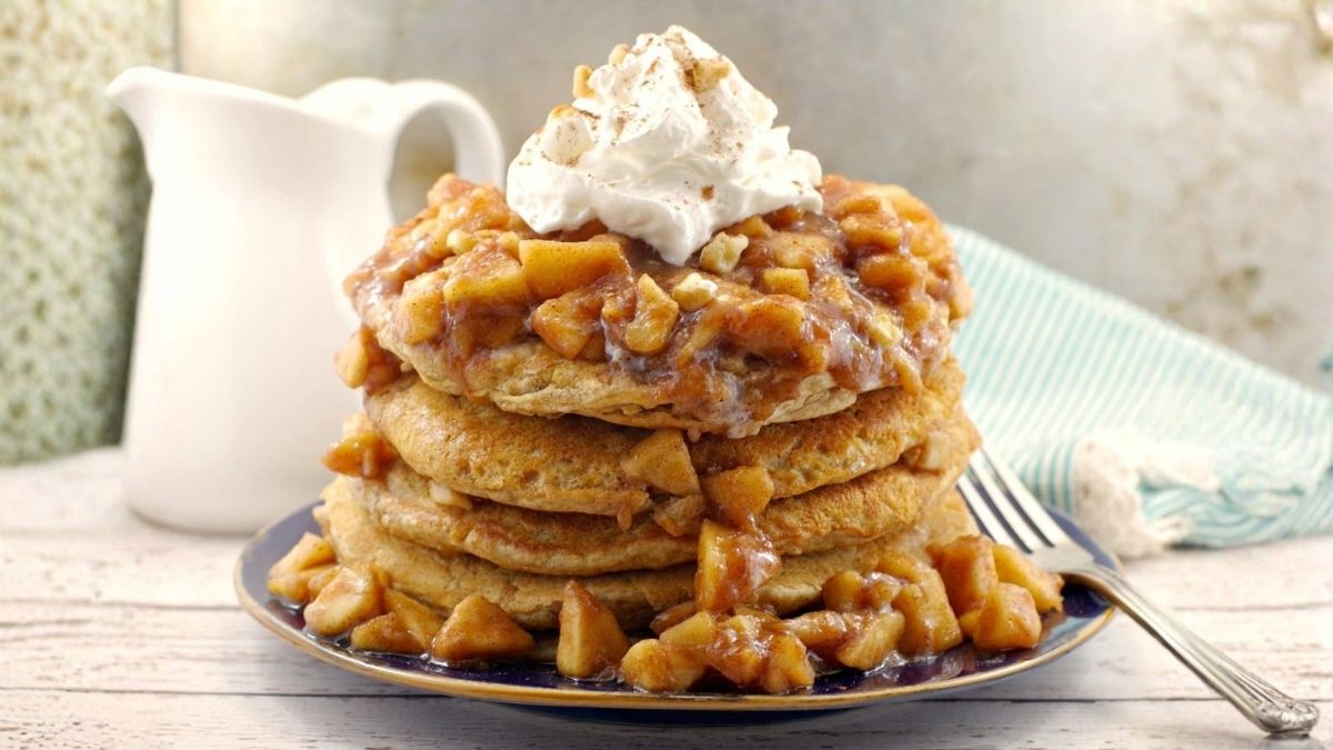Stack of harvest grain and nut pancakes topped with warm cinnamon apples, creamy glaze, and whipped cream on a breakfast plate.