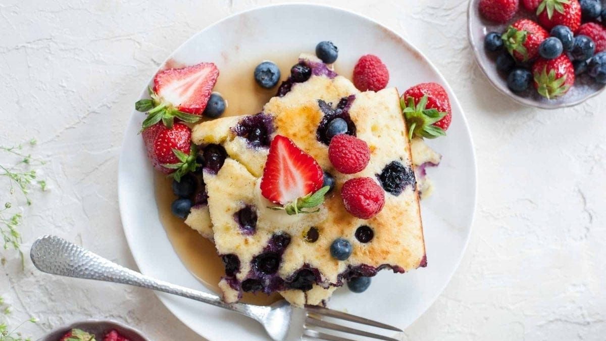 Slice of lemon blueberry sheet pan pancake topped with fresh strawberries, raspberries, and blueberries, drizzled with syrup on a white plate.