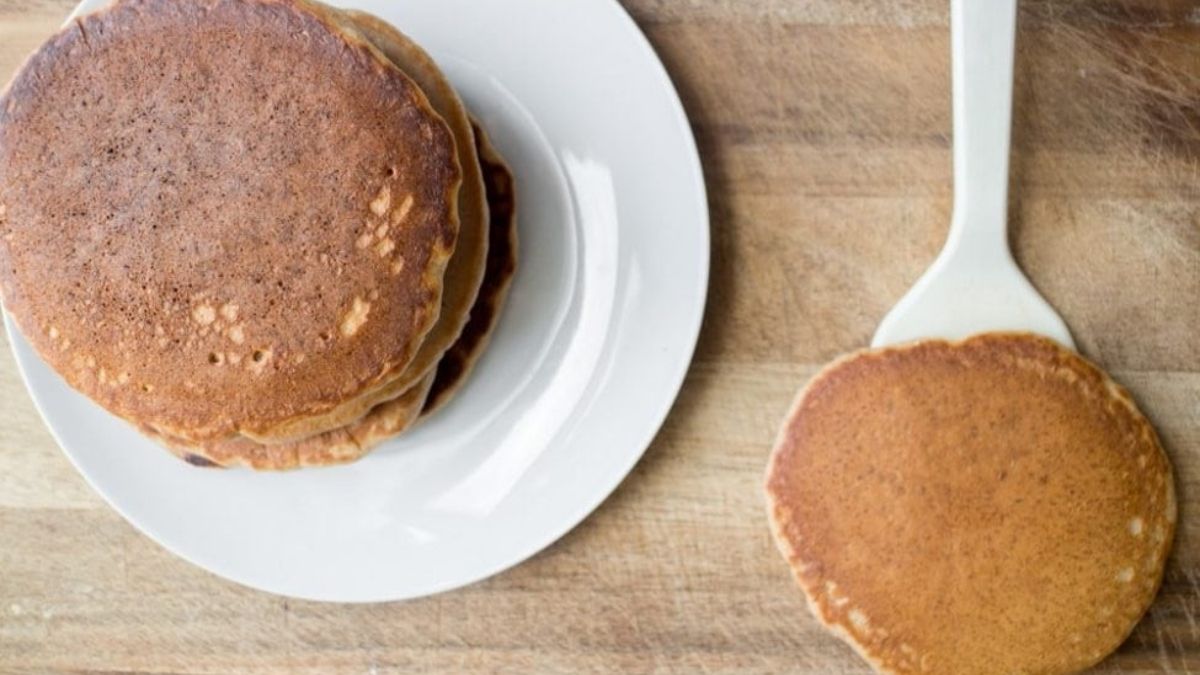 Stack of homemade pancakes on a white plate with a freshly cooked pancake on a spatula beside them on a wooden countertop.