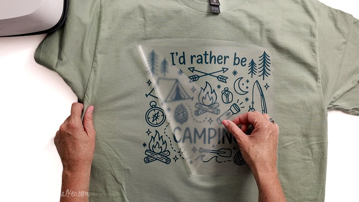 Hands peeling the clear carrier sheet from an “I’d Rather Be Camping” iron-on vinyl design after heat pressing it onto a light green T-shirt.