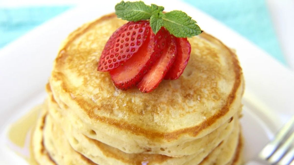 Stack of fluffy homemade pancakes topped with sliced strawberries, fresh mint leaves, and maple syrup on a white plate.