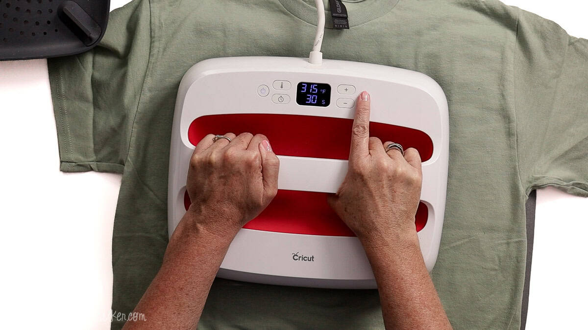 Hands using a Cricut EasyPress to apply an iron-on vinyl design to a light green T-shirt, with the heat press set to 315°F for 30 seconds.