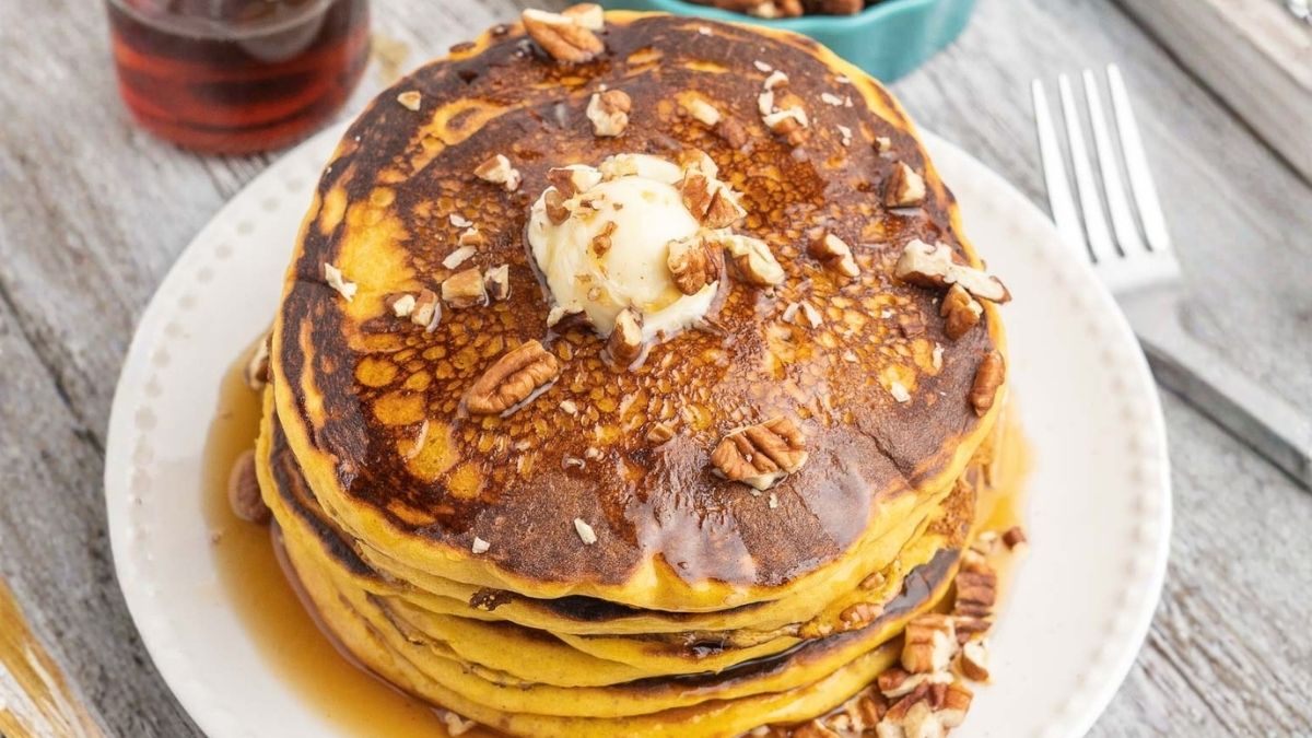 Stack of pumpkin pancakes topped with butter, chopped pecans, and maple syrup on a white plate over a rustic wooden table.