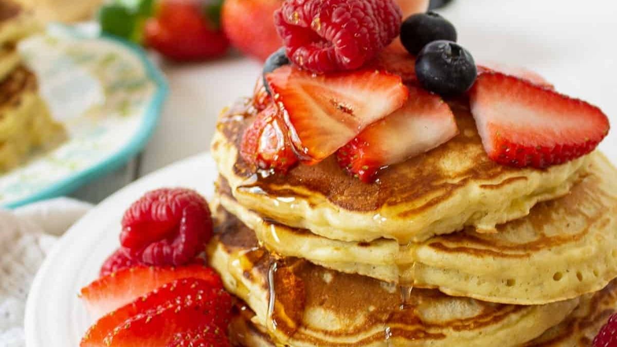 Stack of fluffy quinoa pancakes topped with sliced strawberries, raspberries, blueberries, and maple syrup on a white plate.