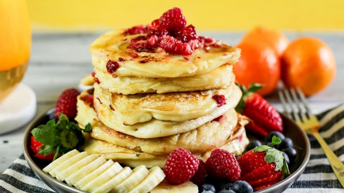 Stack of fluffy berry pancakes topped with raspberries, served with fresh strawberries, blueberries, banana slices, and citrus fruit on a plate.