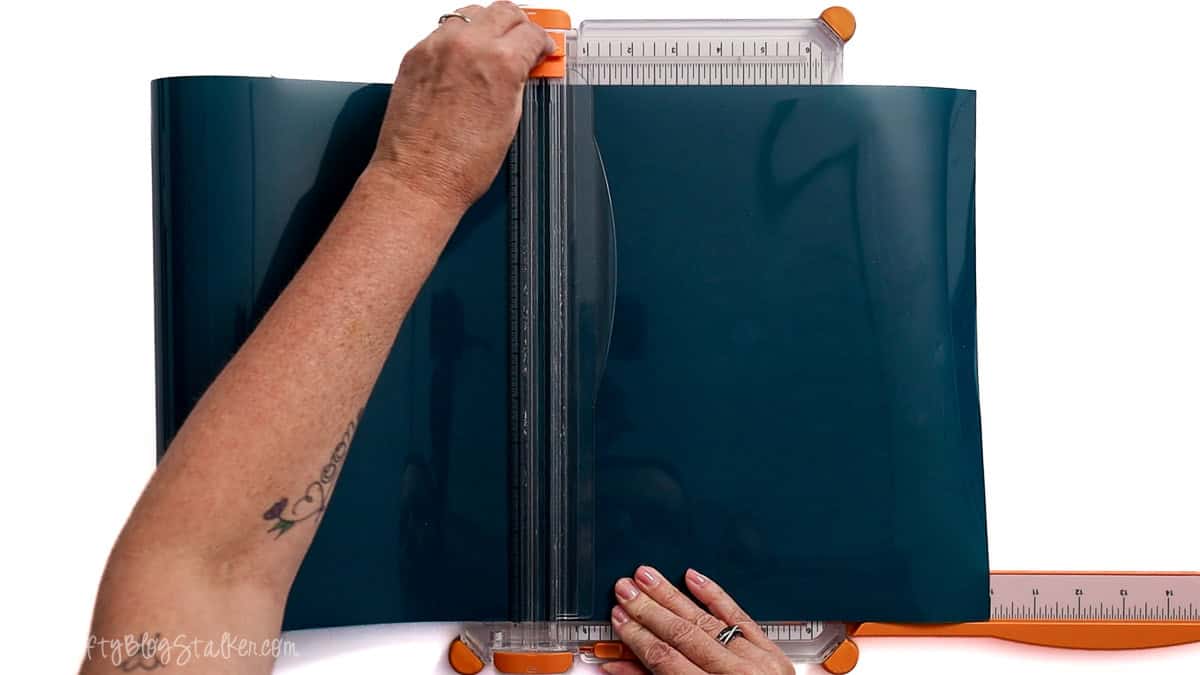 Hands using a paper trimmer to cut a sheet of dark blue iron-on vinyl, preparing material for a Cricut crafting project.
