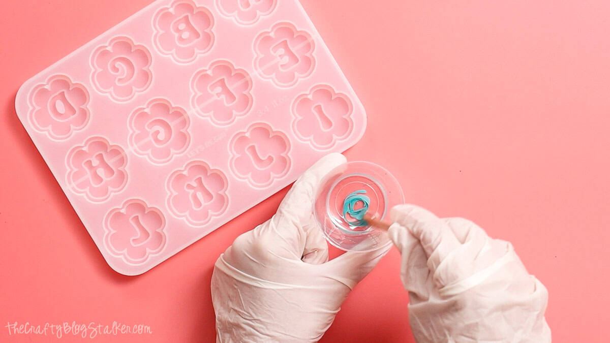Gloved hands mixing turquoise resin pigment into clear epoxy resin in a small measuring cup beside a flower-shaped silicone keychain mold.