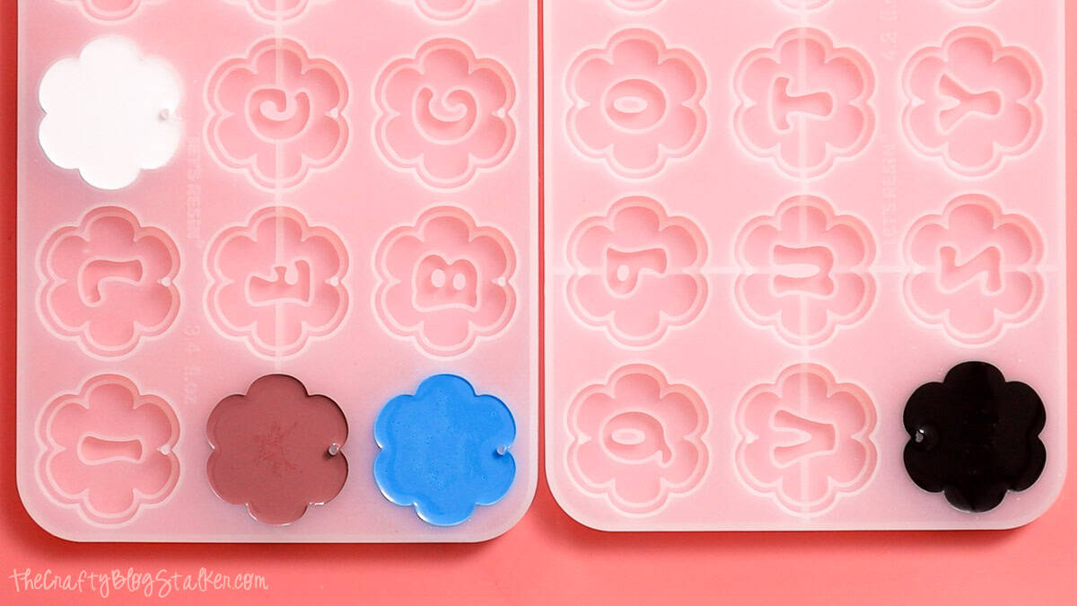 Flower-shaped silicone molds filled with white, blue, brown, and black resin curing to create personalized alphabet resin keychains.