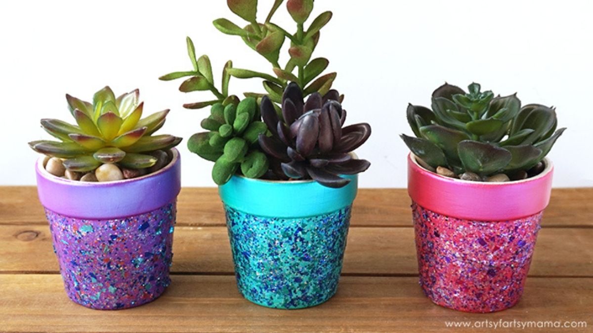 Three small succulent plants in colorful glitter flower pots painted in purple, blue, and pink sit on a wooden tabletop.