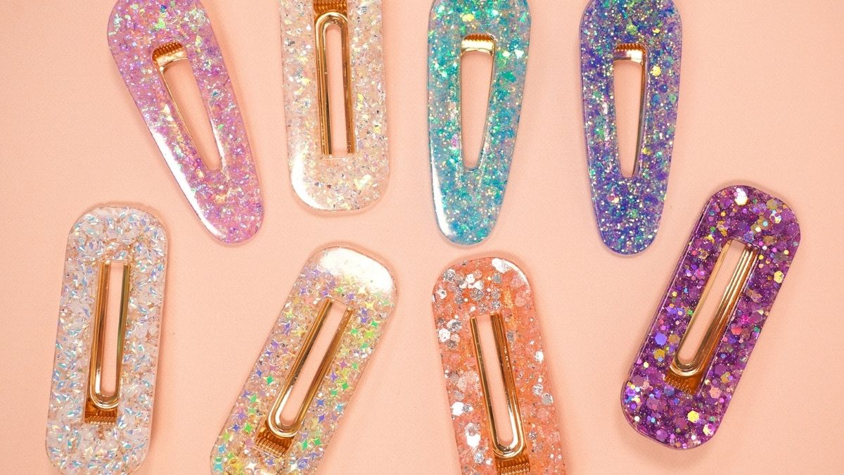Colorful handmade resin hair clips with glitter and iridescent flakes in pink, purple, blue, white, and peach arranged on a peach background.