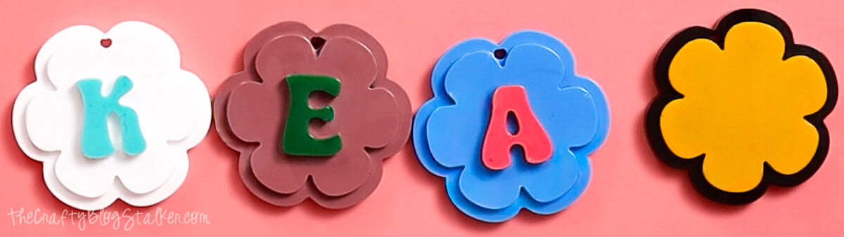 Four flower-shaped resin keychain pieces in white, brown, blue, and yellow with colorful letters displayed on a pink background.