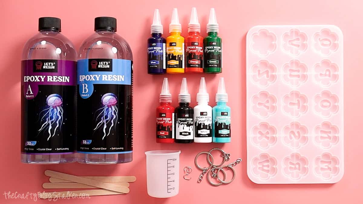 Flat lay of resin keychain supplies including epoxy resin, pigment colors, flower mold, mixing cup, craft sticks, and metal key rings on a pink background.