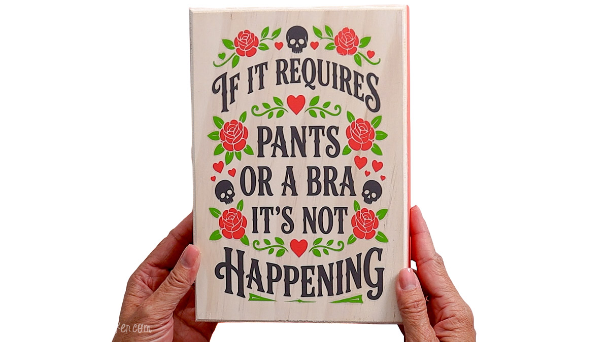 Hands hold a whitewashed wood sign with black text, red roses, green leaves, hearts, and skulls that reads, If It Requires Pants or a Bra It's Not Happening