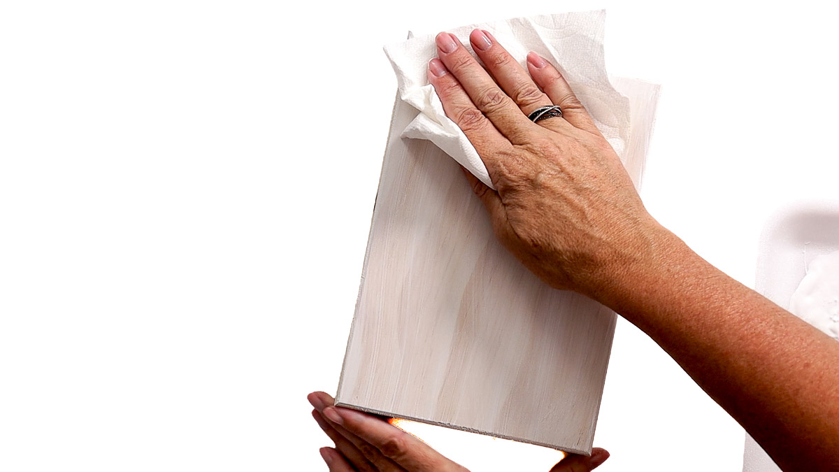 Hand wiping excess whitewash from a wood sign blank with a paper towel to create a light, rustic wood finish.