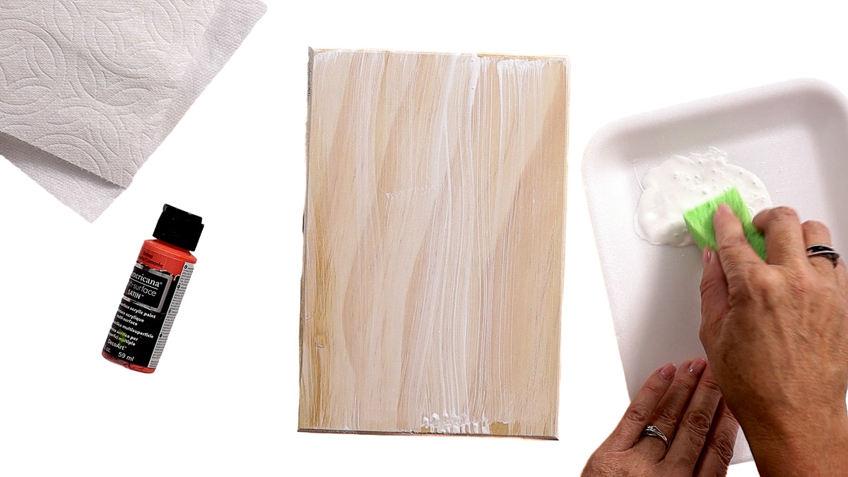 Applying a whitewash to a wood sign blank with white acrylic paint and a sponge, with paint, paper towels, and supplies nearby.