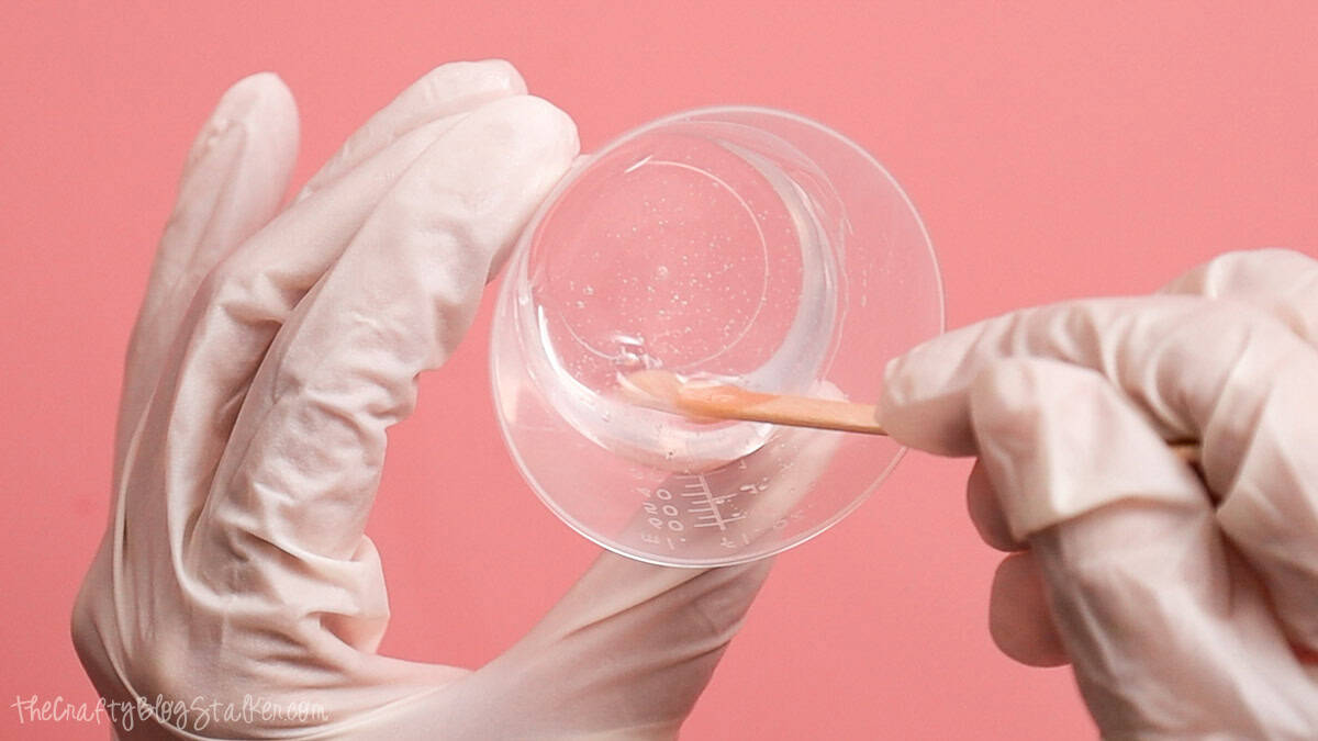 Gloved hands stirring clear mixed epoxy resin in a measuring cup with a wooden craft stick on a pink work surface.