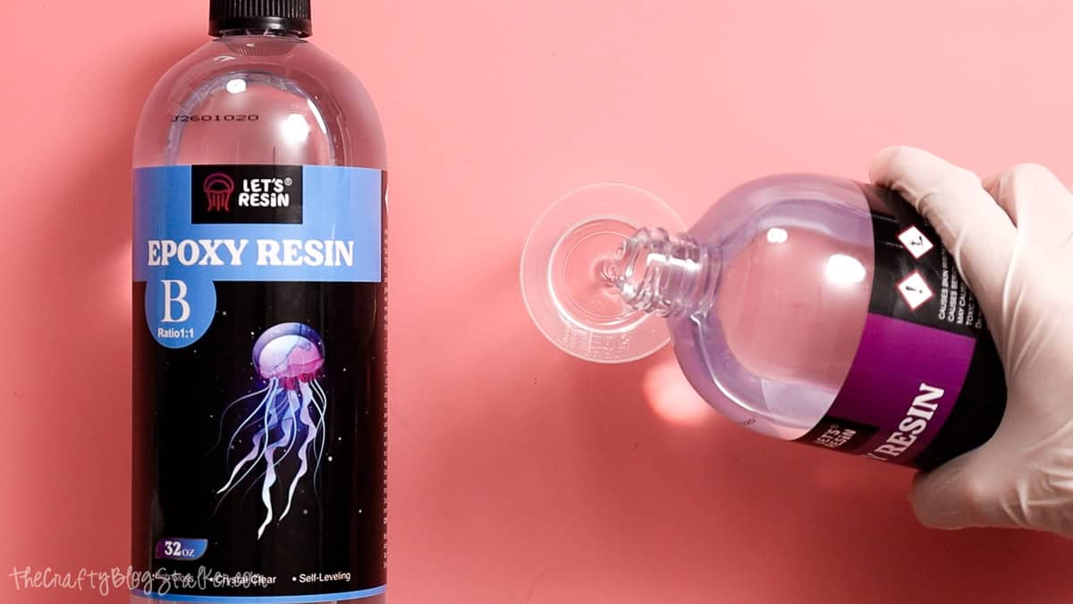 Hand pouring epoxy resin into a mixing cup beside bottles of Let's Resin epoxy resin on a pink work surface.