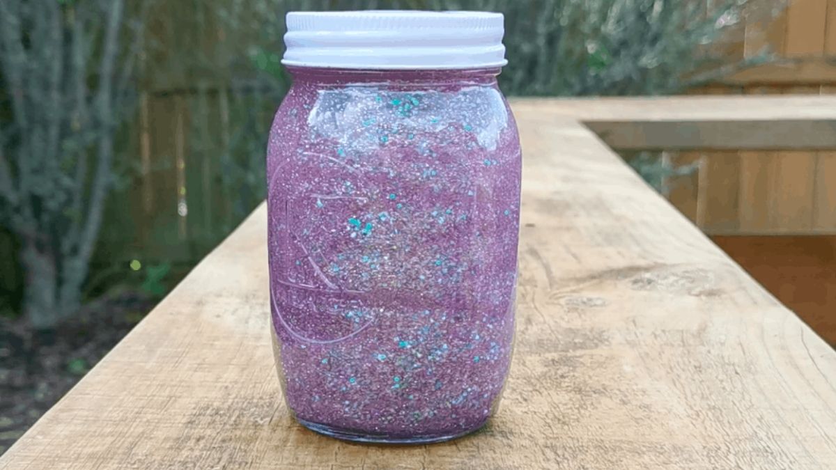 Purple glitter sensory bottle made from a mason jar with silver and blue glitter suspended inside, displayed outdoors on a wooden table.