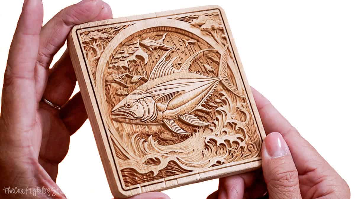 Hands holding a square wooden coaster engraved with a detailed fish and underwater scene, showcasing intricate laser engraving on natural wood.