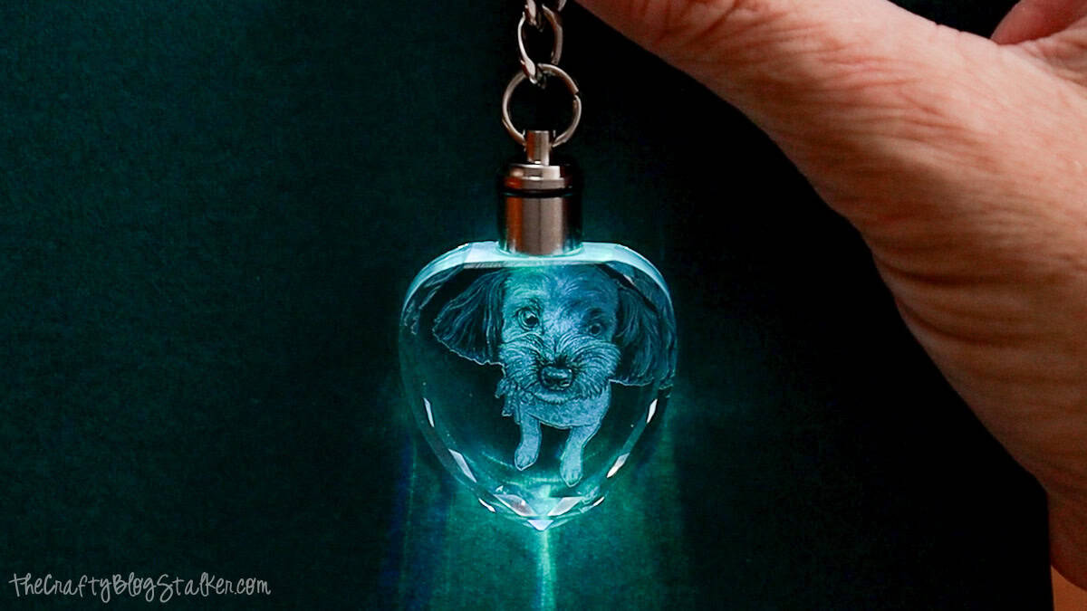 Heart-shaped crystal keychain with a 3D laser-engraved dog portrait, held in a hand against a dark background with glowing blue light.
