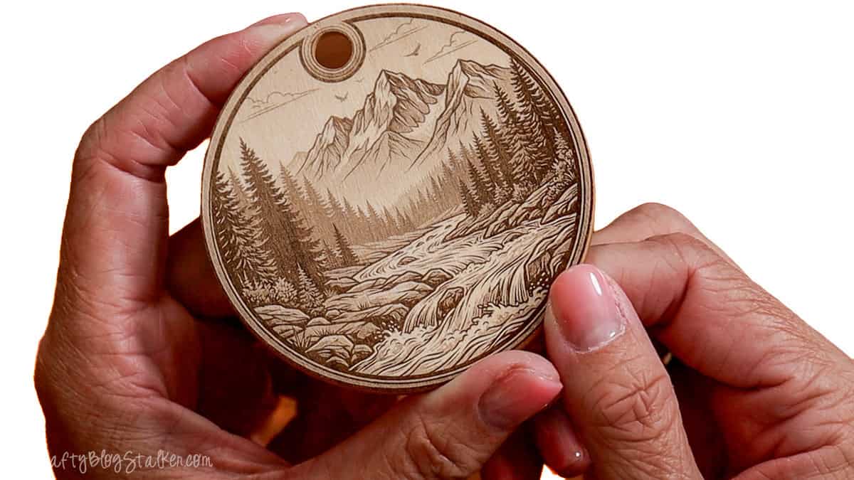 Hands holding a round wooden ornament engraved with a detailed mountain landscape, pine trees, and a flowing river, showcasing fine laser engraving.