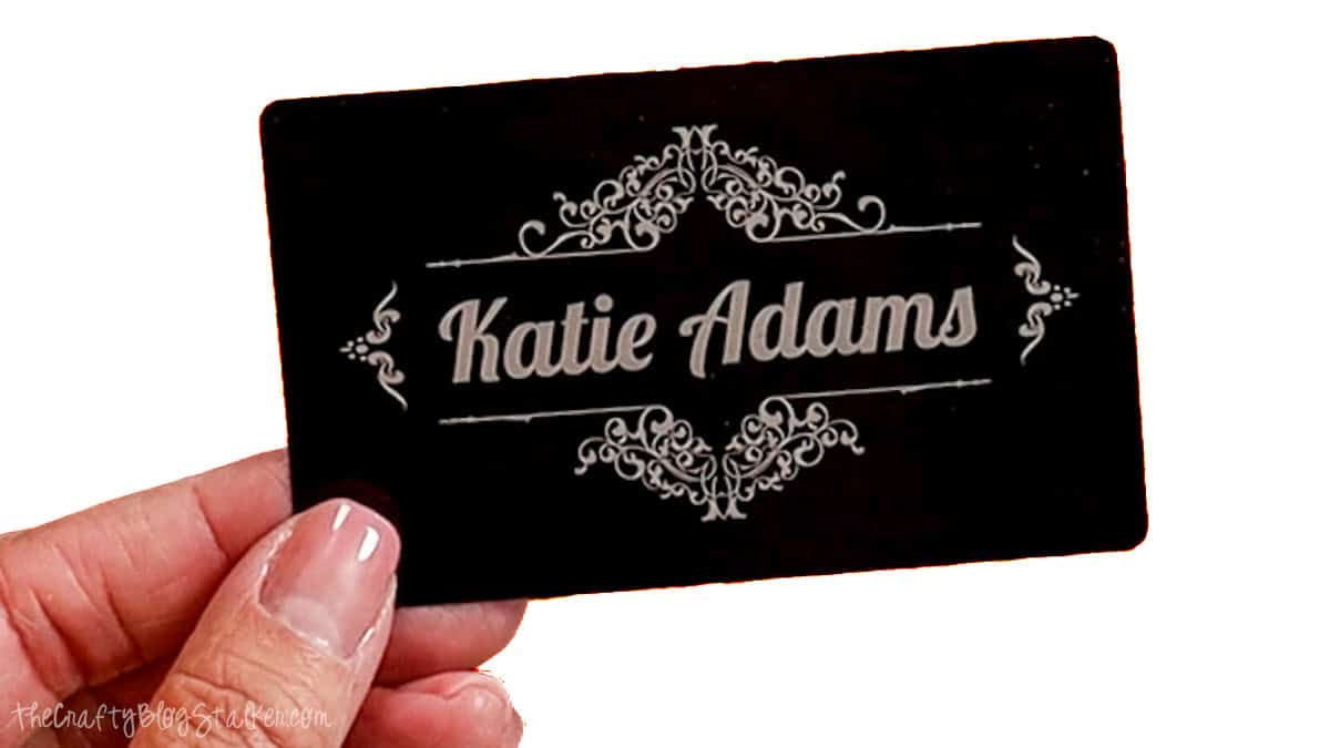 Hand holding a black metal business card engraved with the name Katie Adams and decorative scrollwork, showcasing crisp laser engraving.