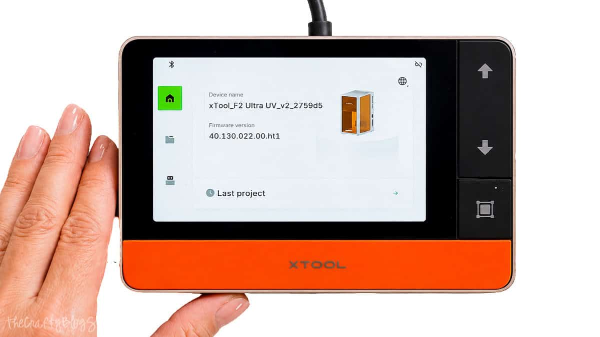 Hand holding the xTool F2 Ultra touchscreen controller displaying the device dashboard, firmware version, and machine status screen.
