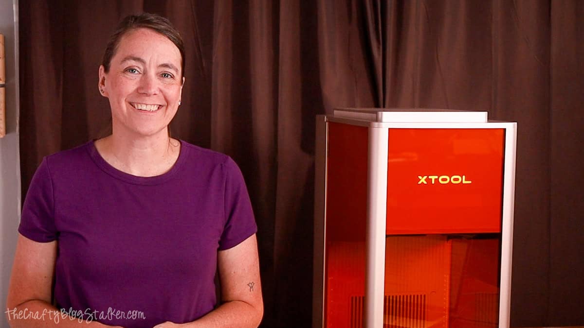 Woman standing beside an xTool F2 Ultra laser cutter, introducing an unboxing and review in a home craft studio.