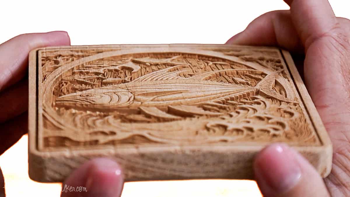 Hands holding a thick wooden coaster engraved with a detailed fish and ocean scene, highlighting the depth and precision of laser engraving.