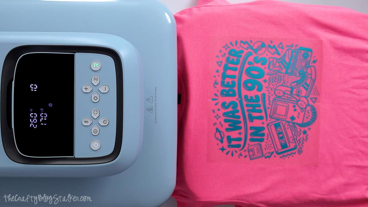 Blue heat press beside a pink T-shirt with a teal 90s puff vinyl design positioned on the front for pressing.