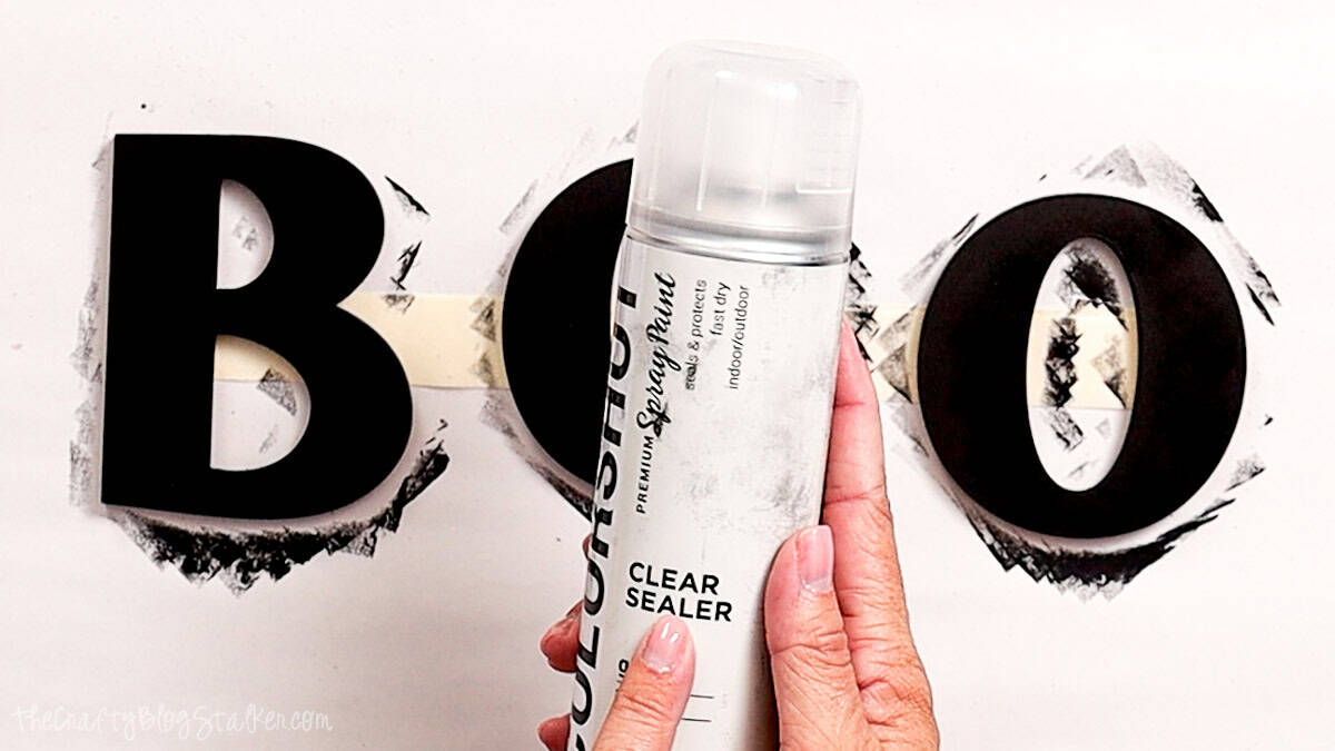 Hand holding a can of clear spray sealer in front of freshly painted black wooden BOO letters for a Halloween door hanger.