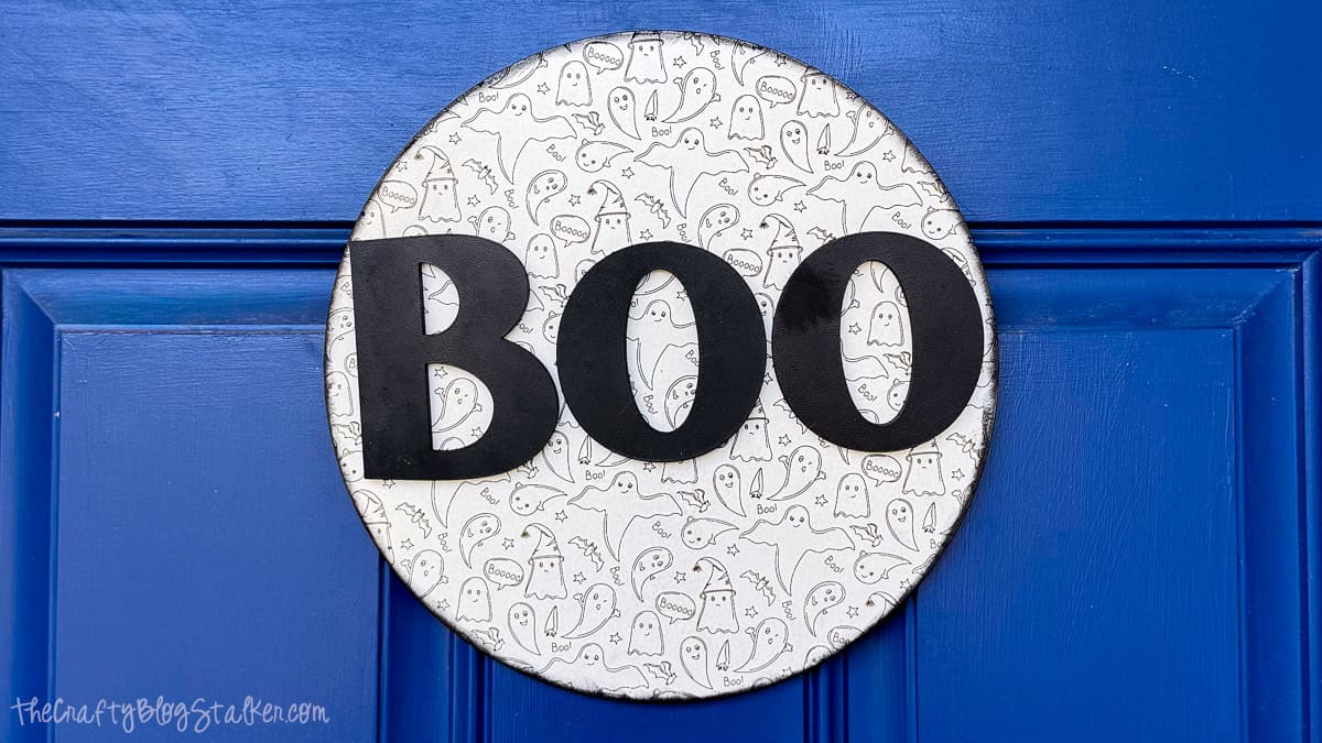 Finished round Halloween BOO door hanger with black letters and an engraved ghost pattern displayed on a bright blue front door.