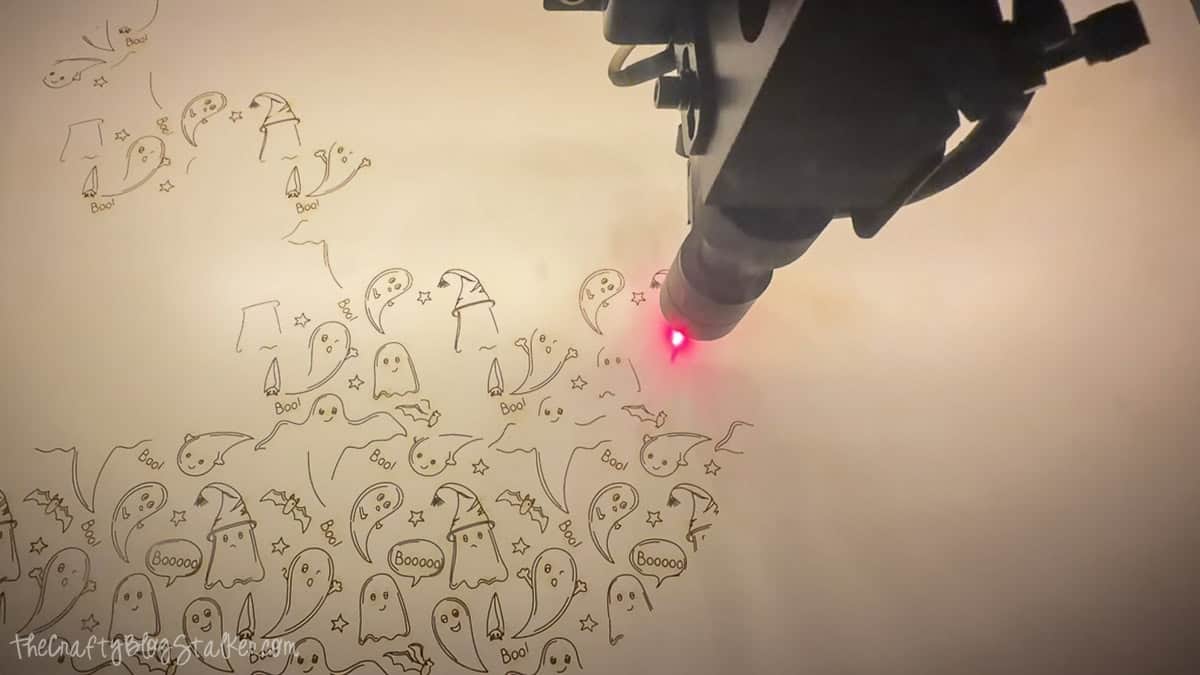 Laser engraver etching a repeating Halloween ghost, bat, star, and witch hat pattern onto wood for a DIY Boo door hanger.