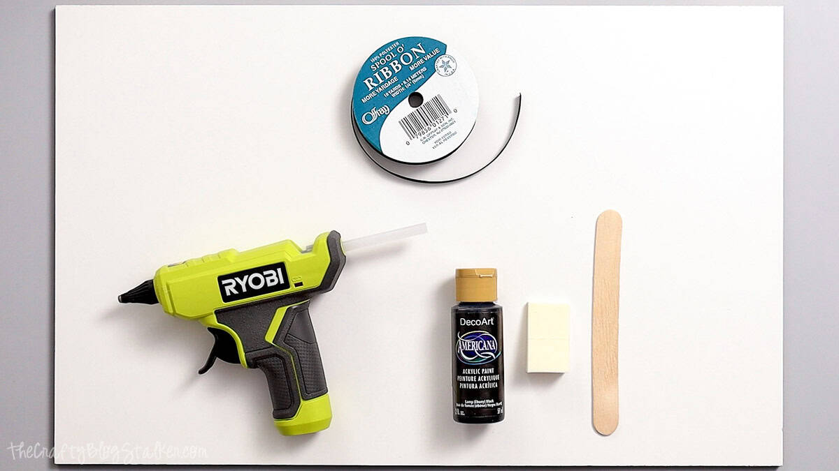Craft supplies for a Halloween door hanger including a hot glue gun, black ribbon, black acrylic paint, foam brush, and wooden craft stick.