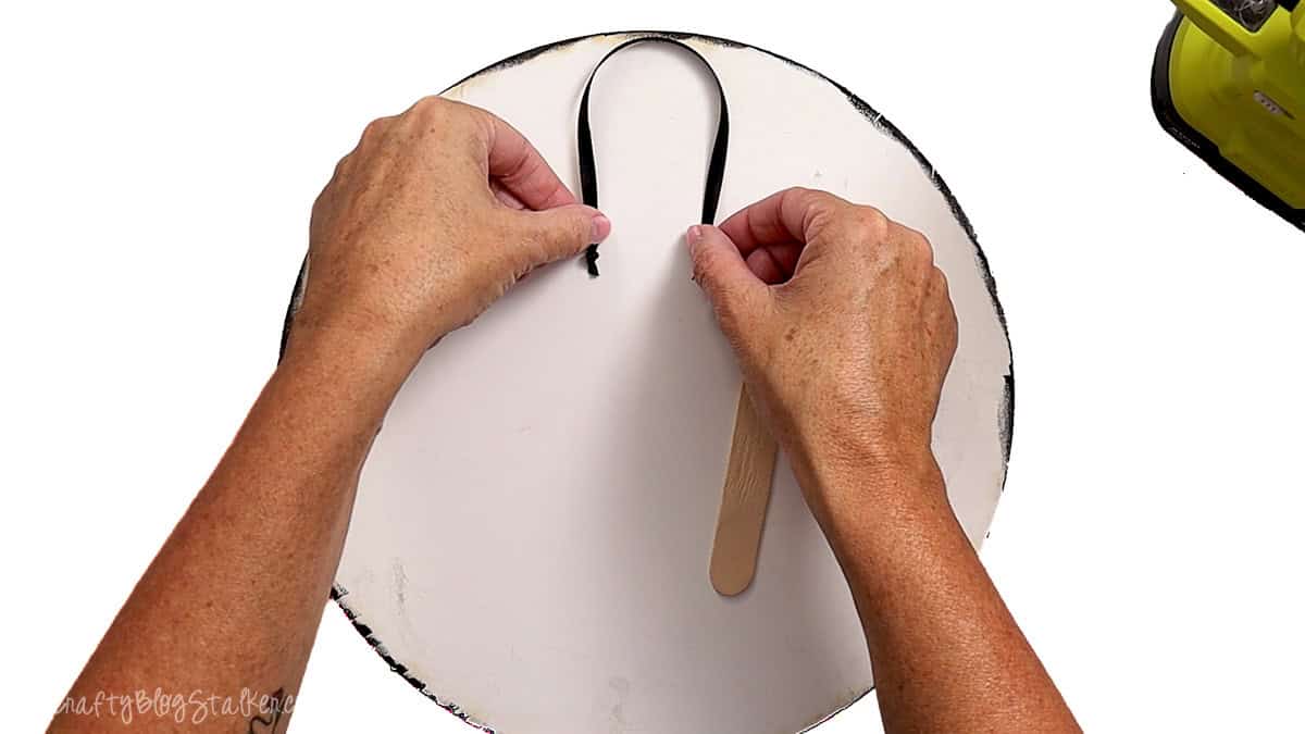 Hands attaching a black ribbon loop to the back of a round wooden Halloween door hanger for hanging.