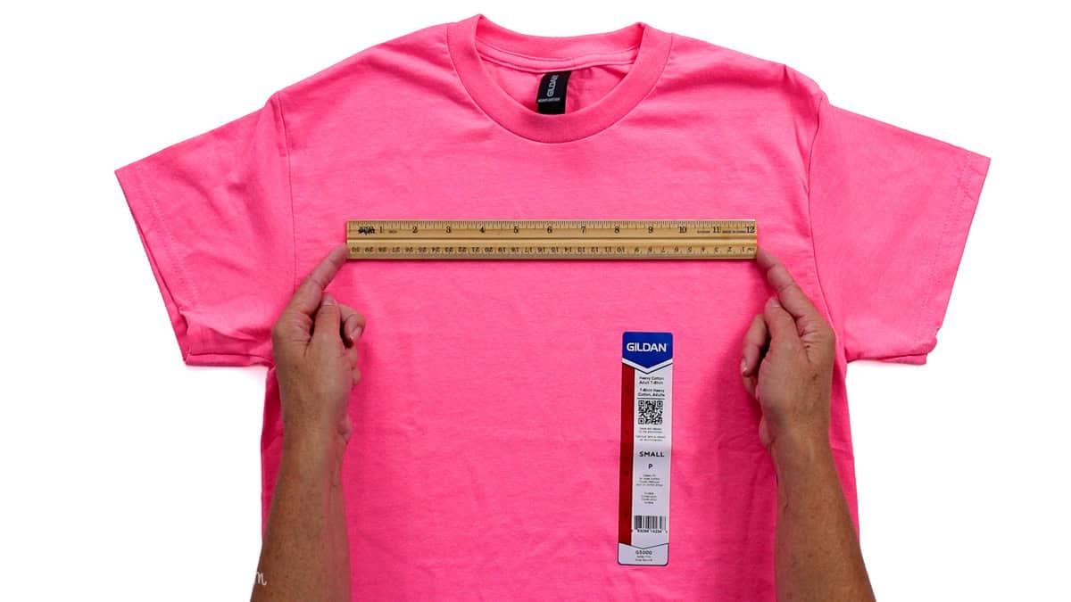 Hands measuring the width of a pink T-shirt with a wooden ruler to determine placement for a puff vinyl design.