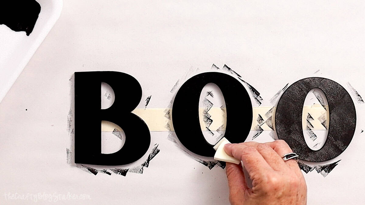 Hand painting wooden BOO letters black with a foam brush for a DIY Halloween door hanger.