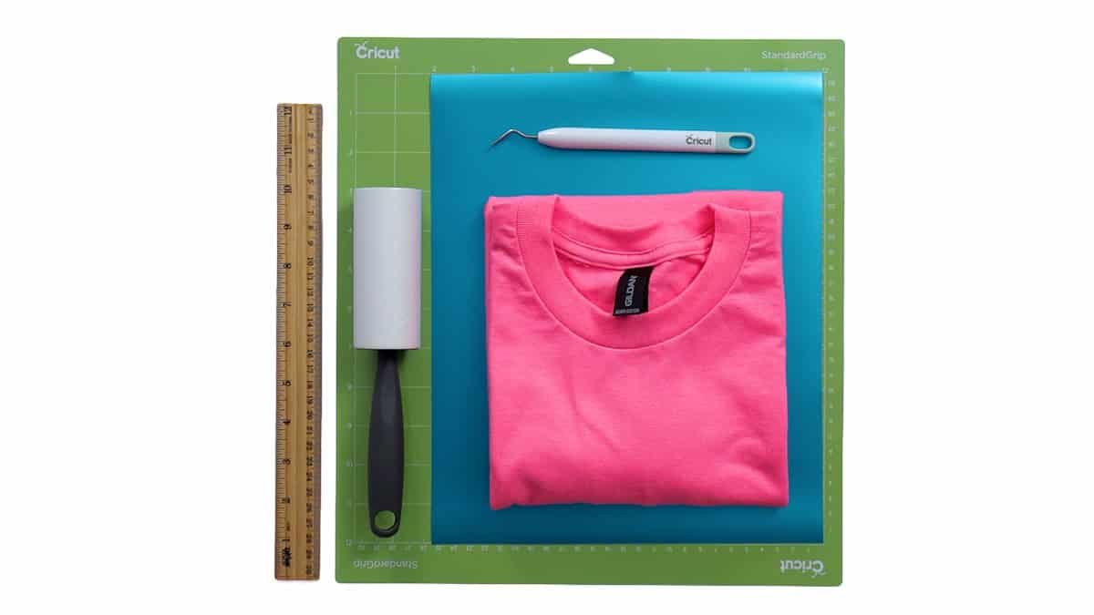 Supplies for a puff vinyl T-shirt project including a pink shirt, Cricut mat, blue vinyl, weeding tool, lint roller, and ruler.