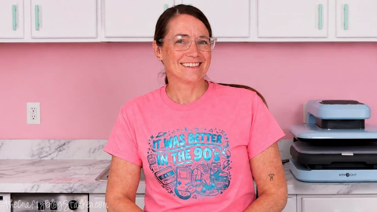 Smiling woman wearing a pink T-shirt with a teal It Was Better in the 90s puff vinyl design, standing beside a heat press.