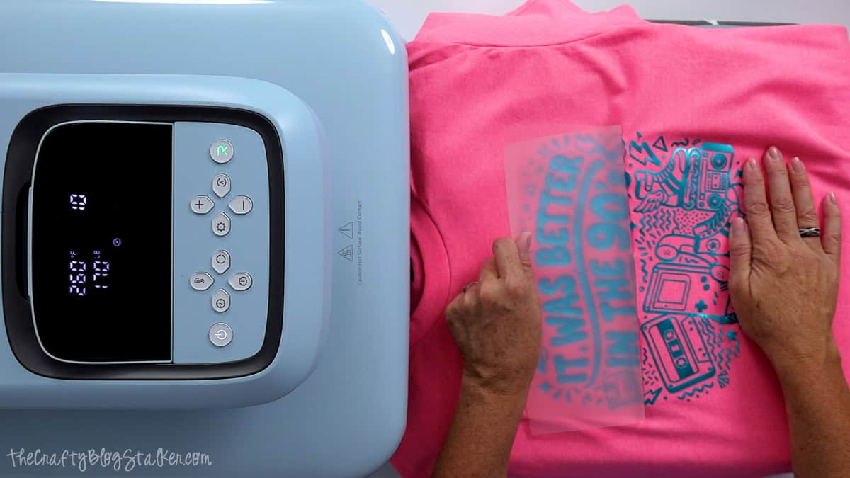 Hands peeling the clear carrier sheet from a teal puff vinyl design after heat pressing it onto a pink T-shirt.