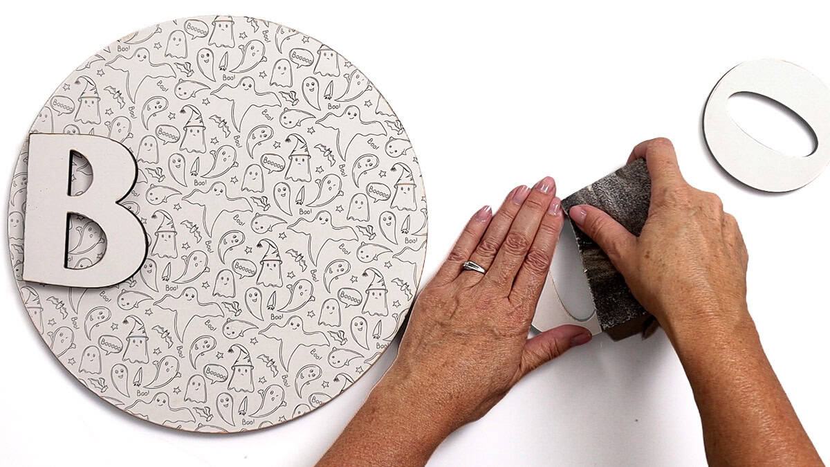 Hands sanding the edge of a wooden O beside a round ghost-patterned Halloween sign and BOO letter pieces.