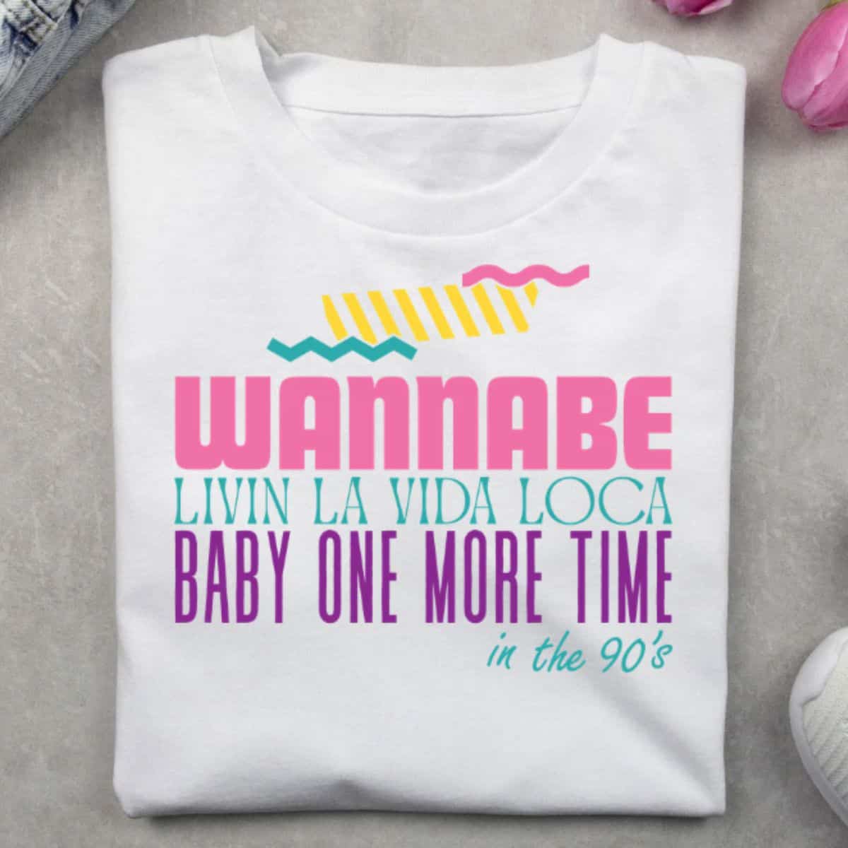 Folded white T-shirt with colorful 90s-inspired text reading Wannabe, Livin' La Vida Loca, Baby One More Time, and in the 90s.